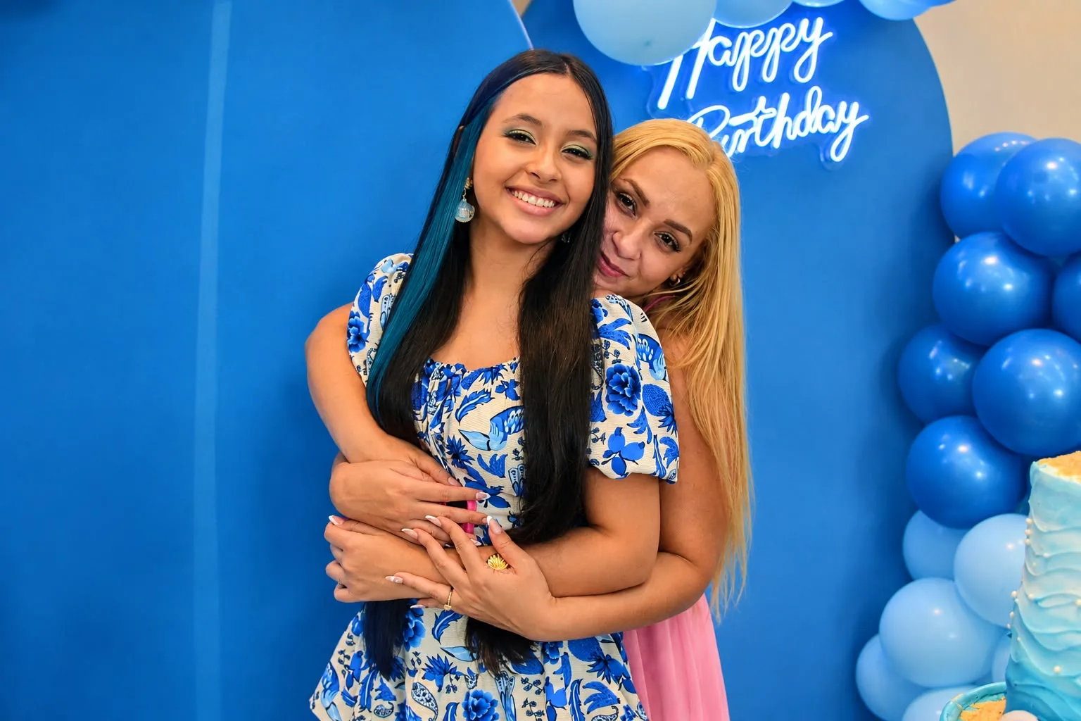 Duas mulheres abraçadas diante da decoração azul da festa de 15 anos